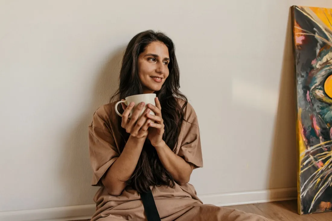 A woman sitting against a wall at home, smiling and holding a mug with both hands.